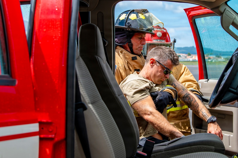 Two firefighters in turnout gear are visible through the open door of a red fire vehicle. One firefighter wearing a black helmet is assisting or extracting another individual from the vehicle cab. The scene appears to be a rescue operation or training exercise, possibly involving vehicle extrication.