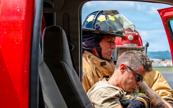 Two firefighters in turnout gear are visible through the open door of a red fire vehicle. One firefighter wearing a black helmet is assisting or extracting another individual from the vehicle cab. The scene appears to be a rescue operation or training exercise, possibly involving vehicle extrication.