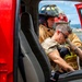 Two firefighters in turnout gear are visible through the open door of a red fire vehicle. One firefighter wearing a black helmet is assisting or extracting another individual from the vehicle cab. The scene appears to be a rescue operation or training exercise, possibly involving vehicle extrication.