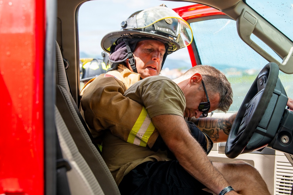 A firefighter in full turnout gear and helmet is reaching into a vehicle through the open driver's side door, appearing to assist or extract a person slumped over the steering wheel. The scene appears to depict an emergency rescue scenario, likely a vehicle accident response or training exercise. The firefighter is wearing a helmet marked 'FIRE DEPT' and is actively engaged with the incapacitated individual.