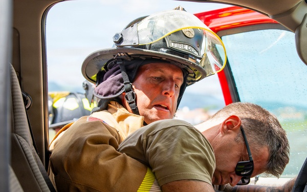 A firefighter in full turnout gear and helmet is reaching into a vehicle through the open driver's side door, appearing to assist or extract a person slumped over the steering wheel. The scene appears to depict an emergency rescue scenario, likely a vehicle accident response or training exercise. The firefighter is wearing a helmet marked 'FIRE DEPT' and is actively engaged with the incapacitated individual.