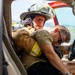 A firefighter in full turnout gear and helmet is reaching into a vehicle through the open driver's side door, appearing to assist or extract a person slumped over the steering wheel. The scene appears to depict an emergency rescue scenario, likely a vehicle accident response or training exercise. The firefighter is wearing a helmet marked 'FIRE DEPT' and is actively engaged with the incapacitated individual.