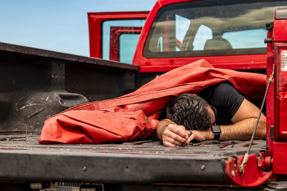 A person is lying face-down in the bed of a red pickup truck, partially covered by a large red tarp or canvas material. The truck appears to be a U.S. Government vehicle based on the partially visible license plate text. The individual is wearing a black short-sleeve shirt and a smartwatch, with their head resting on their crossed arms.