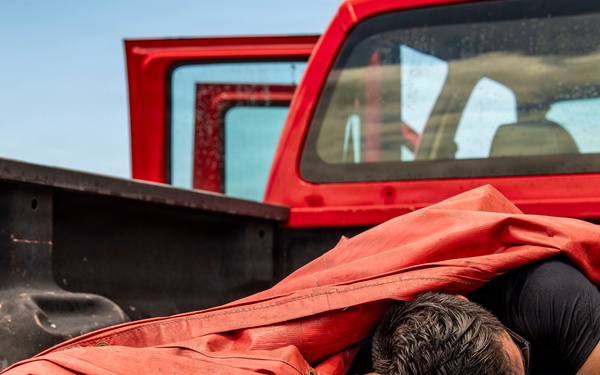 A person is lying face-down in the bed of a red pickup truck, partially covered by a large red tarp or canvas material. The truck appears to be a U.S. Government vehicle based on the partially visible license plate text. The individual is wearing a black short-sleeve shirt and a smartwatch, with their head resting on their crossed arms.