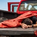 A person is lying face-down in the bed of a red pickup truck, partially covered by a large red tarp or canvas material. The truck appears to be a U.S. Government vehicle based on the partially visible license plate text. The individual is wearing a black short-sleeve shirt and a smartwatch, with their head resting on their crossed arms.
