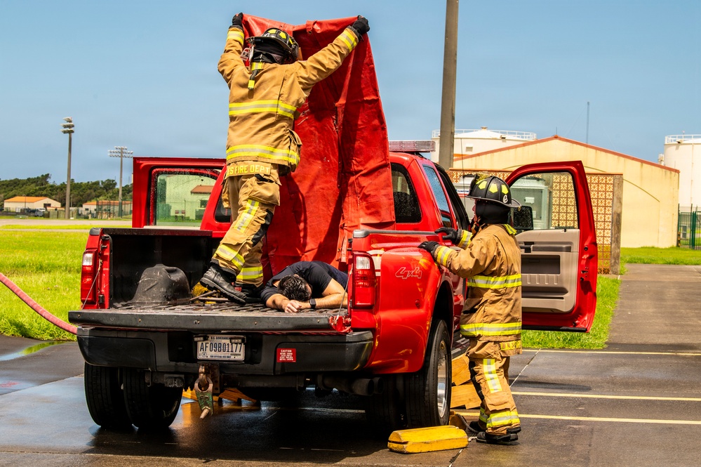 Two firefighters in full turnout gear are conducting what appears to be a rescue training exercise involving a red 4x4 pickup truck. One firefighter is standing in the truck bed holding a red tarp overhead, while a simulated victim lies face-down in the truck bed. A second firefighter stands beside the open rear door, assisting with the operation.