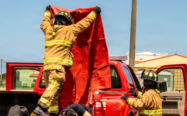 Two firefighters in full turnout gear are conducting what appears to be a rescue training exercise involving a red 4x4 pickup truck. One firefighter is standing in the truck bed holding a red tarp overhead, while a simulated victim lies face-down in the truck bed. A second firefighter stands beside the open rear door, assisting with the operation.