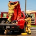 Two firefighters in full turnout gear are conducting what appears to be a rescue training exercise involving a red 4x4 pickup truck. One firefighter is standing in the truck bed holding a red tarp overhead, while a simulated victim lies face-down in the truck bed. A second firefighter stands beside the open rear door, assisting with the operation.