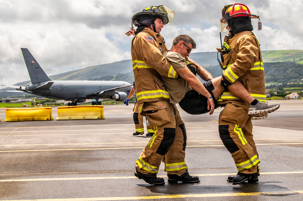 Two firefighters in full turnout gear with high-visibility yellow striping are carrying an incapacitated person across a wet airport tarmac. A large military or civilian jet aircraft is visible in the background, suggesting this is likely an emergency response drill or actual incident at an airfield. The setting appears to be a coastal or island location based on the green hills and wind turbines visible in the background.