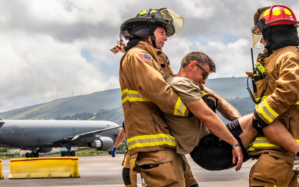 Two firefighters in full turnout gear with high-visibility yellow striping are carrying an incapacitated person across a wet airport tarmac. A large military or civilian jet aircraft is visible in the background, suggesting this is likely an emergency response drill or actual incident at an airfield. The setting appears to be a coastal or island location based on the green hills and wind turbines visible in the background.