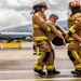 Two firefighters in full turnout gear with high-visibility yellow striping are carrying an incapacitated person across a wet airport tarmac. A large military or civilian jet aircraft is visible in the background, suggesting this is likely an emergency response drill or actual incident at an airfield. The setting appears to be a coastal or island location based on the green hills and wind turbines visible in the background.