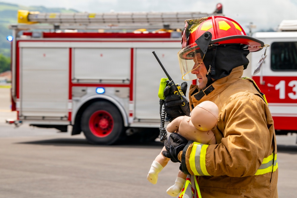 A firefighter in full turnout gear, wearing a red helmet with face shield, is holding a baby training mannequin while communicating on a handheld radio. A red fire truck with active blue emergency lights is visible in the background, suggesting this is a training exercise or drill. The setting appears to be an open tarmac or staging area.