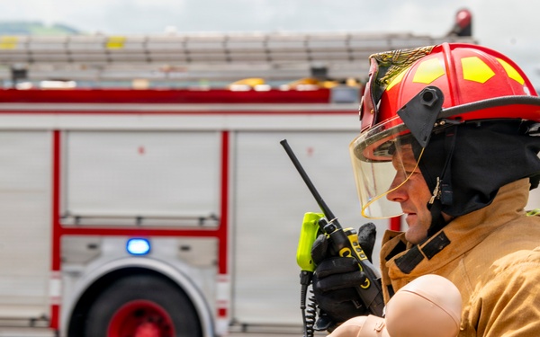 A firefighter in full turnout gear, wearing a red helmet with face shield, is holding a baby training mannequin while communicating on a handheld radio. A red fire truck with active blue emergency lights is visible in the background, suggesting this is a training exercise or drill. The setting appears to be an open tarmac or staging area.