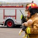 A firefighter in full turnout gear, wearing a red helmet with face shield, is holding a baby training mannequin while communicating on a handheld radio. A red fire truck with active blue emergency lights is visible in the background, suggesting this is a training exercise or drill. The setting appears to be an open tarmac or staging area.