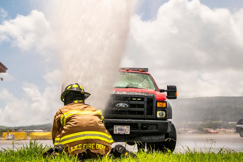 A firefighter from the Lajes Fire Department is crouched on grass in full turnout gear, observing a powerful water geyser erupting in front of a Ford F-series U.S. Government vehicle (plate: AF09B01177). The scene appears to be a training exercise or a ruptured water line on what looks like an airfield or tarmac. The water spray is drenching the truck and surrounding area under a partly cloudy sky.