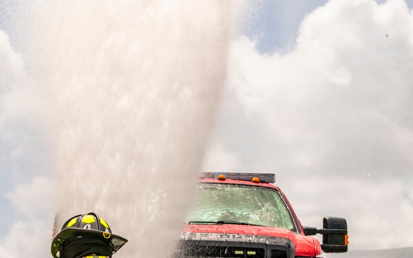 A firefighter from the Lajes Fire Department is crouched on grass in full turnout gear, observing a powerful water geyser erupting in front of a Ford F-series U.S. Government vehicle (plate: AF09B01177). The scene appears to be a training exercise or a ruptured water line on what looks like an airfield or tarmac. The water spray is drenching the truck and surrounding area under a partly cloudy sky.