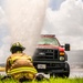 A firefighter from the Lajes Fire Department is crouched on grass in full turnout gear, observing a powerful water geyser erupting in front of a Ford F-series U.S. Government vehicle (plate: AF09B01177). The scene appears to be a training exercise or a ruptured water line on what looks like an airfield or tarmac. The water spray is drenching the truck and surrounding area under a partly cloudy sky.