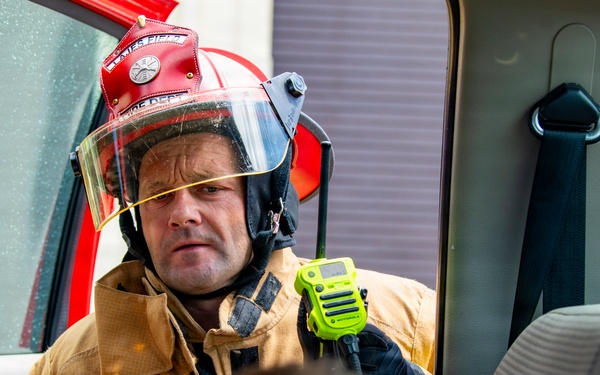 A firefighter in full gear, including a red helmet with face shield and tan turnout jacket, is leaning into an open vehicle door attending to a person inside. The firefighter is holding a bright yellow-green radio/walkie-talkie and appears to be assessing or communicating during what looks like an emergency response or rescue scenario. A second person, visible only from the back of the head, is positioned inside the vehicle.