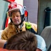A firefighter in full gear, including a red helmet with face shield and tan turnout jacket, is leaning into an open vehicle door attending to a person inside. The firefighter is holding a bright yellow-green radio/walkie-talkie and appears to be assessing or communicating during what looks like an emergency response or rescue scenario. A second person, visible only from the back of the head, is positioned inside the vehicle.