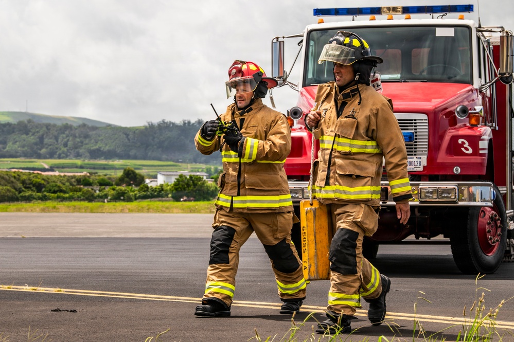 Two firefighters in full turnout gear are walking on a paved surface, likely a tarmac or airfield, in front of a red fire truck numbered '3'. One firefighter is holding a radio/walkie-talkie while the other carries a yellow equipment case. The background shows green hills and an overcast sky, suggesting a rural or semi-rural location.