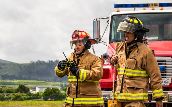 Two firefighters in full turnout gear are walking on a paved surface, likely a tarmac or airfield, in front of a red fire truck numbered '3'. One firefighter is holding a radio/walkie-talkie while the other carries a yellow equipment case. The background shows green hills and an overcast sky, suggesting a rural or semi-rural location.