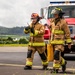 Two firefighters in full turnout gear are walking on a paved surface, likely a tarmac or airfield, in front of a red fire truck numbered '3'. One firefighter is holding a radio/walkie-talkie while the other carries a yellow equipment case. The background shows green hills and an overcast sky, suggesting a rural or semi-rural location.