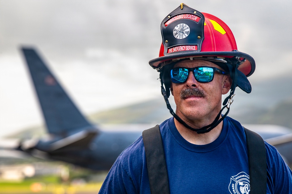 A male firefighter wearing a red helmet and blue mirrored sunglasses stands in the foreground, looking slightly off-camera with a subtle expression. His helmet reads 'FIRE EMERGENCY SERVICE' and his t-shirt displays 'CRASH' and 'RICKENBACKER' text. In the blurred background, a large military or commercial aircraft tail section is visible under an overcast sky.