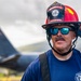 A male firefighter wearing a red helmet and blue mirrored sunglasses stands in the foreground, looking slightly off-camera with a subtle expression. His helmet reads 'FIRE EMERGENCY SERVICE' and his t-shirt displays 'CRASH' and 'RICKENBACKER' text. In the blurred background, a large military or commercial aircraft tail section is visible under an overcast sky.