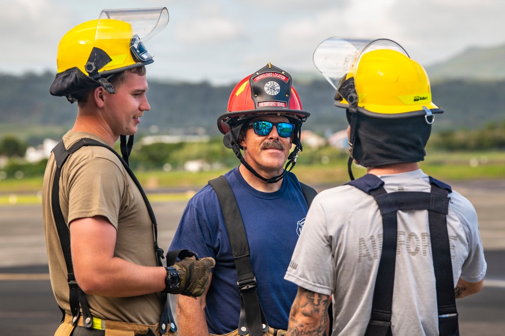 Three firefighters or emergency service personnel are standing on what appears to be an airfield tarmac, engaged in conversation. Two individuals wear yellow firefighting helmets with face shields, while the central figure wears a red fire helmet with blue mirrored sunglasses and a mustache. The setting appears to be a tropical or subtropical location based on the green hills and vegetation visible in the background.