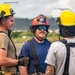 Three firefighters or emergency service personnel are standing on what appears to be an airfield tarmac, engaged in conversation. Two individuals wear yellow firefighting helmets with face shields, while the central figure wears a red fire helmet with blue mirrored sunglasses and a mustache. The setting appears to be a tropical or subtropical location based on the green hills and vegetation visible in the background.