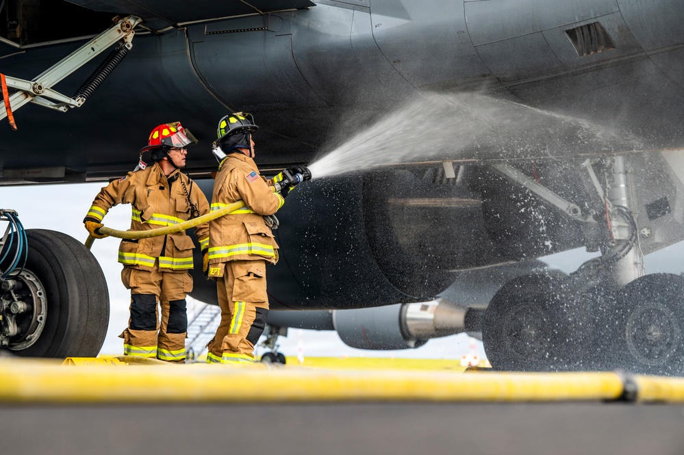 Two firefighters in full turnout gear are directing a water hose at the landing gear or engine nacelle area of a large military aircraft on a tarmac. The scene appears to be a firefighting drill or emergency response exercise at an airfield. One firefighter wears a red helmet and the other a black helmet, both in standard tan/yellow reflective protective gear.