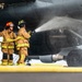 Two firefighters in full turnout gear are directing a water hose at the landing gear or engine nacelle area of a large military aircraft on a tarmac. The scene appears to be a firefighting drill or emergency response exercise at an airfield. One firefighter wears a red helmet and the other a black helmet, both in standard tan/yellow reflective protective gear.