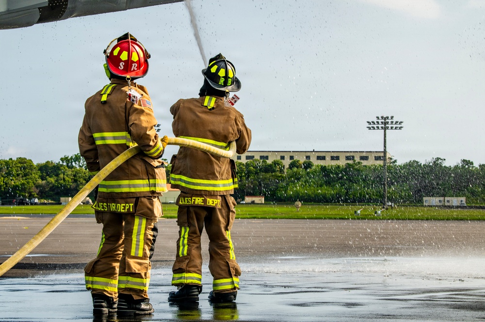 Two firefighters in full turnout gear are operating a fire hose on what appears to be an airport tarmac or airfield. Both are wearing brown protective suits with high-visibility yellow reflective striping and helmets, with water spraying from the hose. The setting suggests a training exercise or emergency response drill at an airfield facility.