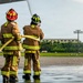 Two firefighters in full turnout gear are operating a fire hose on what appears to be an airport tarmac or airfield. Both are wearing brown protective suits with high-visibility yellow reflective striping and helmets, with water spraying from the hose. The setting suggests a training exercise or emergency response drill at an airfield facility.