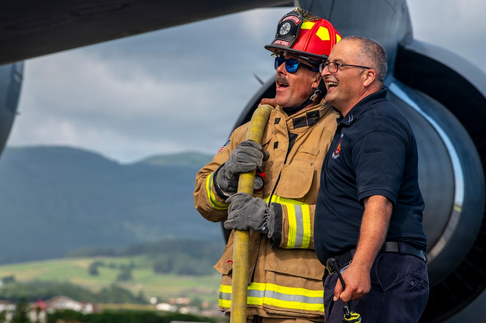 Two firefighters are standing near what appears to be a large aircraft engine or fuselage, both laughing and appearing to share a lighthearted moment. One firefighter is in full turnout gear including a red helmet, blue sunglasses, and gloves while holding a yellow fire hose, while the other wears a navy blue uniform shirt. The background reveals a mountainous, green landscape under overcast skies.