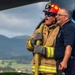 Two firefighters are standing near what appears to be a large aircraft engine or fuselage, both laughing and appearing to share a lighthearted moment. One firefighter is in full turnout gear including a red helmet, blue sunglasses, and gloves while holding a yellow fire hose, while the other wears a navy blue uniform shirt. The background reveals a mountainous, green landscape under overcast skies.