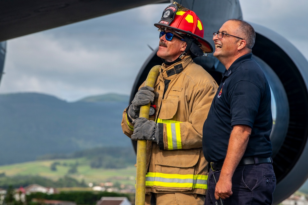 Two men stand in front of a large jet engine, with one dressed in full firefighter turnout gear including a red helmet and carrying a fire hose, and the other in a dark navy uniform shirt. Both appear relaxed and are looking upward, possibly observing aircraft operations. The setting appears to be an airfield with mountainous, green terrain visible in the background.