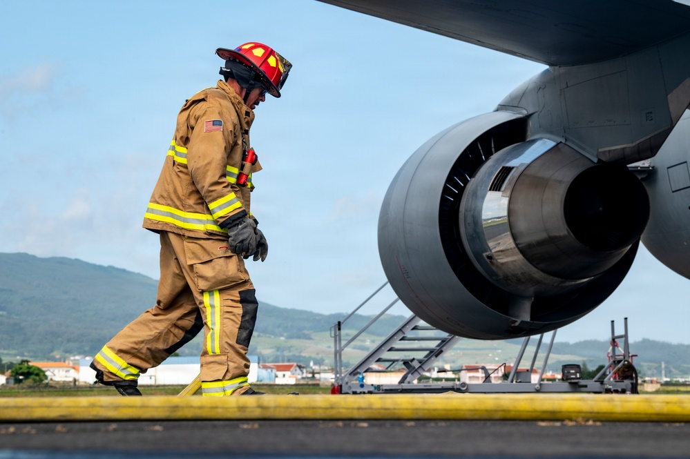 A firefighter in full turnout gear, wearing a red helmet and tan/yellow reflective suit with a US flag patch, walks near a large jet engine on an airport tarmac. The setting appears to be a military or joint-use airfield, with buildings, green hills, and blue skies visible in the background. The scene suggests an aircraft emergency response exercise or inspection activity.