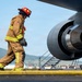 A firefighter in full turnout gear, wearing a red helmet and tan/yellow reflective suit with a US flag patch, walks near a large jet engine on an airport tarmac. The setting appears to be a military or joint-use airfield, with buildings, green hills, and blue skies visible in the background. The scene suggests an aircraft emergency response exercise or inspection activity.