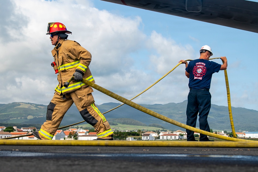 Two firefighters are working together on what appears to be an airport tarmac or runway, handling a large yellow fire hose. One firefighter is in full turnout gear with a red helmet, while the second wears a navy blue shirt labeled 'Bombeiros' (Portuguese for firefighters) and a white hard hat. A residential area and green hills are visible in the background, suggesting a location in Portugal or the Azores.