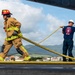 Two firefighters are working together on what appears to be an airport tarmac or runway, handling a large yellow fire hose. One firefighter is in full turnout gear with a red helmet, while the second wears a navy blue shirt labeled 'Bombeiros' (Portuguese for firefighters) and a white hard hat. A residential area and green hills are visible in the background, suggesting a location in Portugal or the Azores.