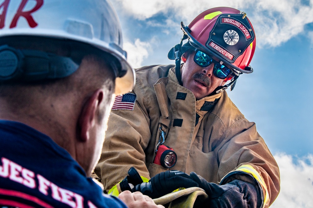 Two firefighters are engaged in a hands-on operational task, likely handling hose equipment or gear during a training exercise or active response. The firefighter in focus wears a red helmet labeled 'UNDISCLOSED LOCATION - FIRE EMERGENCY SERVICE' with an American flag patch on his turnout gear. The image is a close-up, dramatic shot taken from a low angle against a partly cloudy sky.