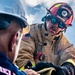 Two firefighters are engaged in a hands-on operational task, likely handling hose equipment or gear during a training exercise or active response. The firefighter in focus wears a red helmet labeled 'UNDISCLOSED LOCATION - FIRE EMERGENCY SERVICE' with an American flag patch on his turnout gear. The image is a close-up, dramatic shot taken from a low angle against a partly cloudy sky.
