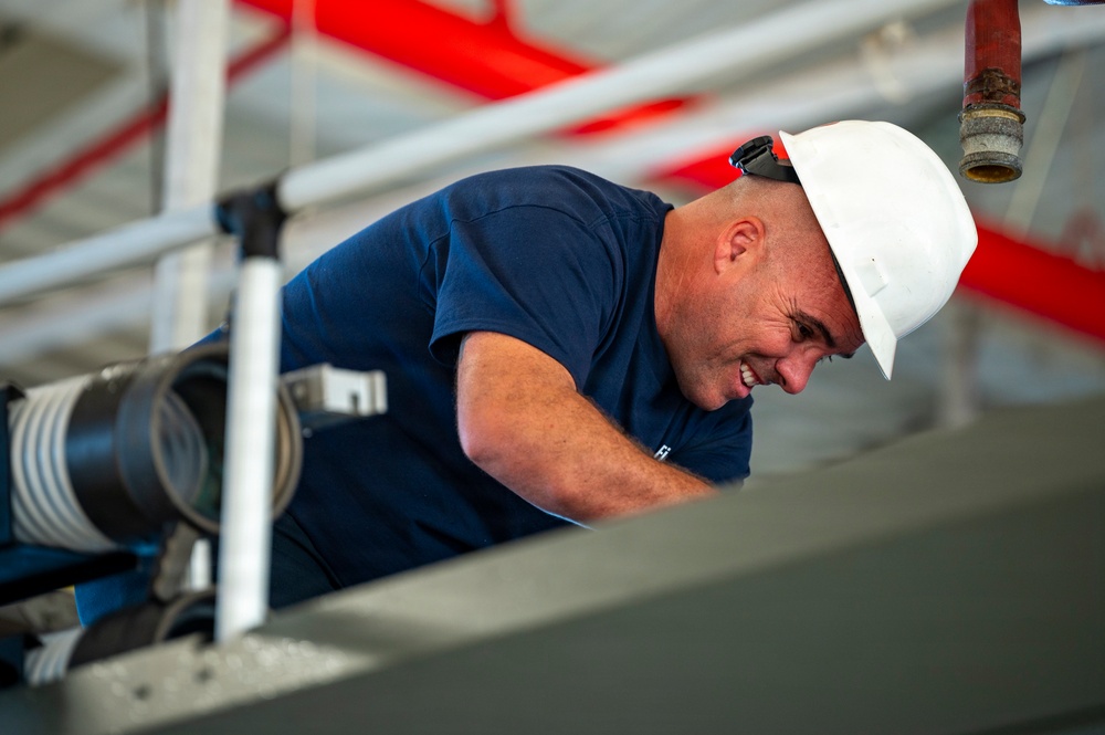 A male worker wearing a white hard hat and navy blue t-shirt is leaning forward and smiling while working in an industrial or construction environment. He appears to be inspecting or working on equipment at an elevated position. The background shows red piping, metal railings, and various industrial infrastructure.