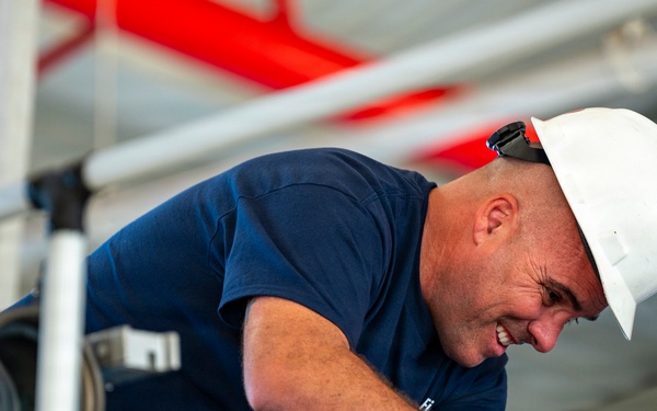 A male worker wearing a white hard hat and navy blue t-shirt is leaning forward and smiling while working in an industrial or construction environment. He appears to be inspecting or working on equipment at an elevated position. The background shows red piping, metal railings, and various industrial infrastructure.