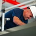 A male worker wearing a white hard hat and navy blue t-shirt is leaning forward and smiling while working in an industrial or construction environment. He appears to be inspecting or working on equipment at an elevated position. The background shows red piping, metal railings, and various industrial infrastructure.