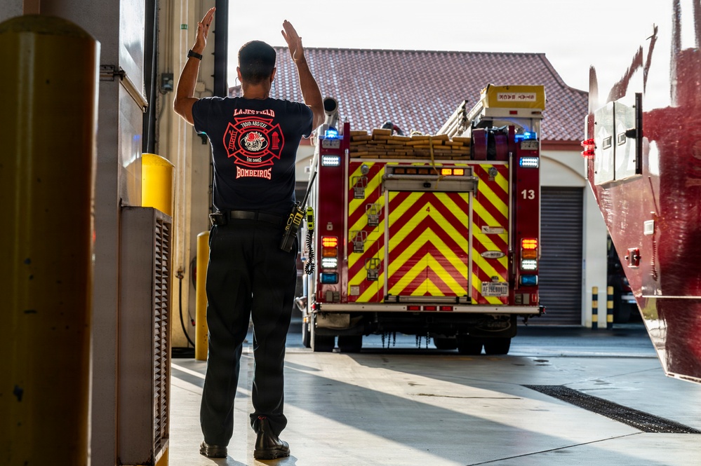 A firefighter from Lajes Field 'Bombeiros' is guiding a fire truck as it backs into a fire station bay. The firefighter has both arms raised, using hand signals to direct the vehicle safely. The fire truck displays prominent red and yellow chevron markings and has its emergency lights activated.