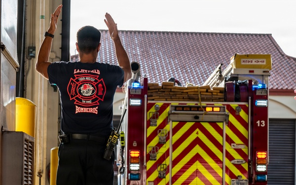 A firefighter from Lajes Field 'Bombeiros' is guiding a fire truck as it backs into a fire station bay. The firefighter has both arms raised, using hand signals to direct the vehicle safely. The fire truck displays prominent red and yellow chevron markings and has its emergency lights activated.