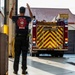 A firefighter from Lajes Field 'Bombeiros' is guiding a fire truck as it backs into a fire station bay. The firefighter has both arms raised, using hand signals to direct the vehicle safely. The fire truck displays prominent red and yellow chevron markings and has its emergency lights activated.