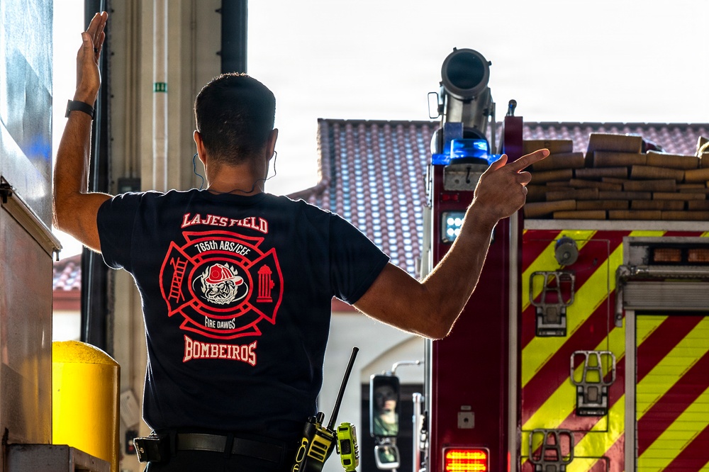 A firefighter wearing a navy blue 'Lajes Field Bombeiros' t-shirt is seen from behind, directing or guiding a fire truck out of a fire station bay. The shirt displays the 765th ABS/CEF unit insignia with a bulldog mascot and the nickname 'Fire Dawgs', indicating a U.S. Air Force fire department unit stationed at Lajes Field in the Azores, Portugal. The firefighter is gesturing with one hand pointing outward while the other is raised against a wall.